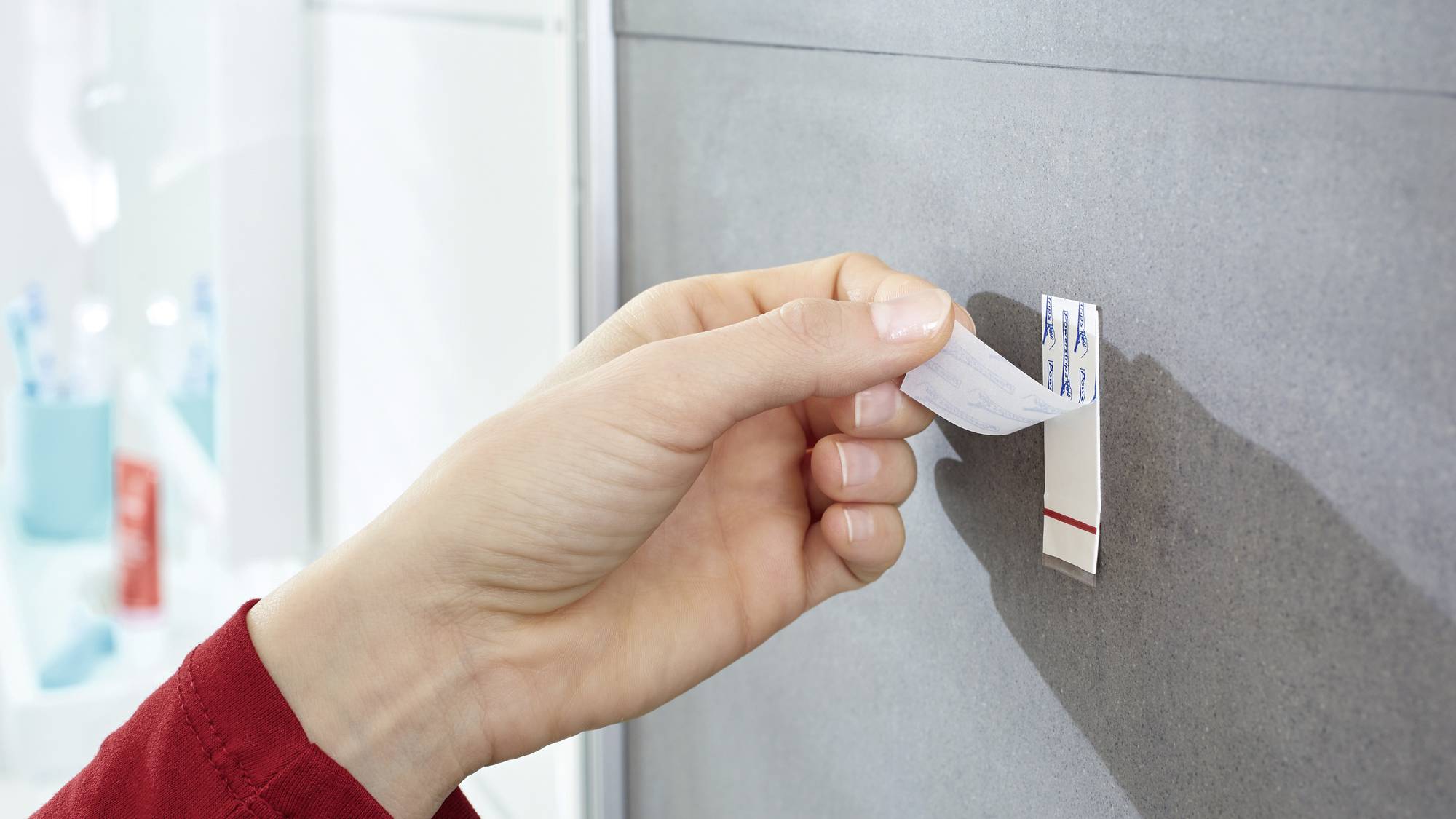 Eine Hand klebt ein Befestigungsband an eine graue Wand. Im Hintergrund sind verschwommen Toilettenartikel sichtbar.