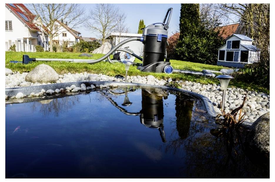 Ein moderner Gartenteich mit klarem Wasser ist von Steinen und Gras umgeben. Ein Teichsauger ruht am Rand und spiegelt sich im Wasser.