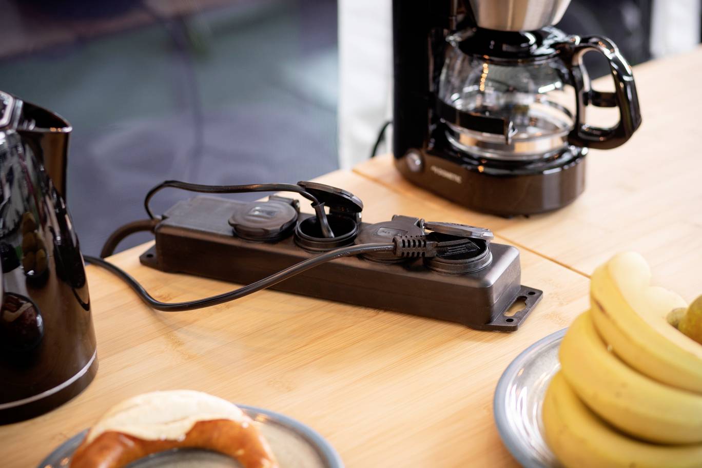 Eine Mehrfachsteckdose auf einem Holztisch verbindet eine Kaffeemaschine und einen Wasserkocher. Daneben liegt ein Teller mit Brezel und Bananen.