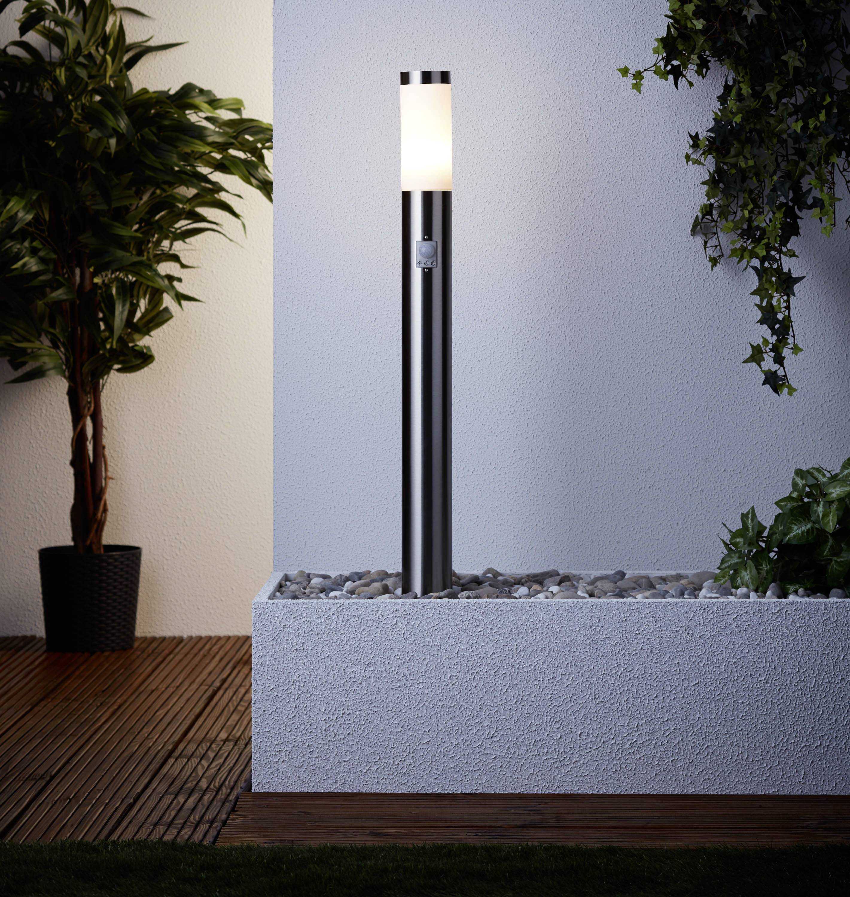 Eine moderne Gartenlampe auf einem Kiesbett, umgeben von Pflanzen auf Holzterrasse im Abendlicht. Beleuchtet eine Ecke eines Gartens.