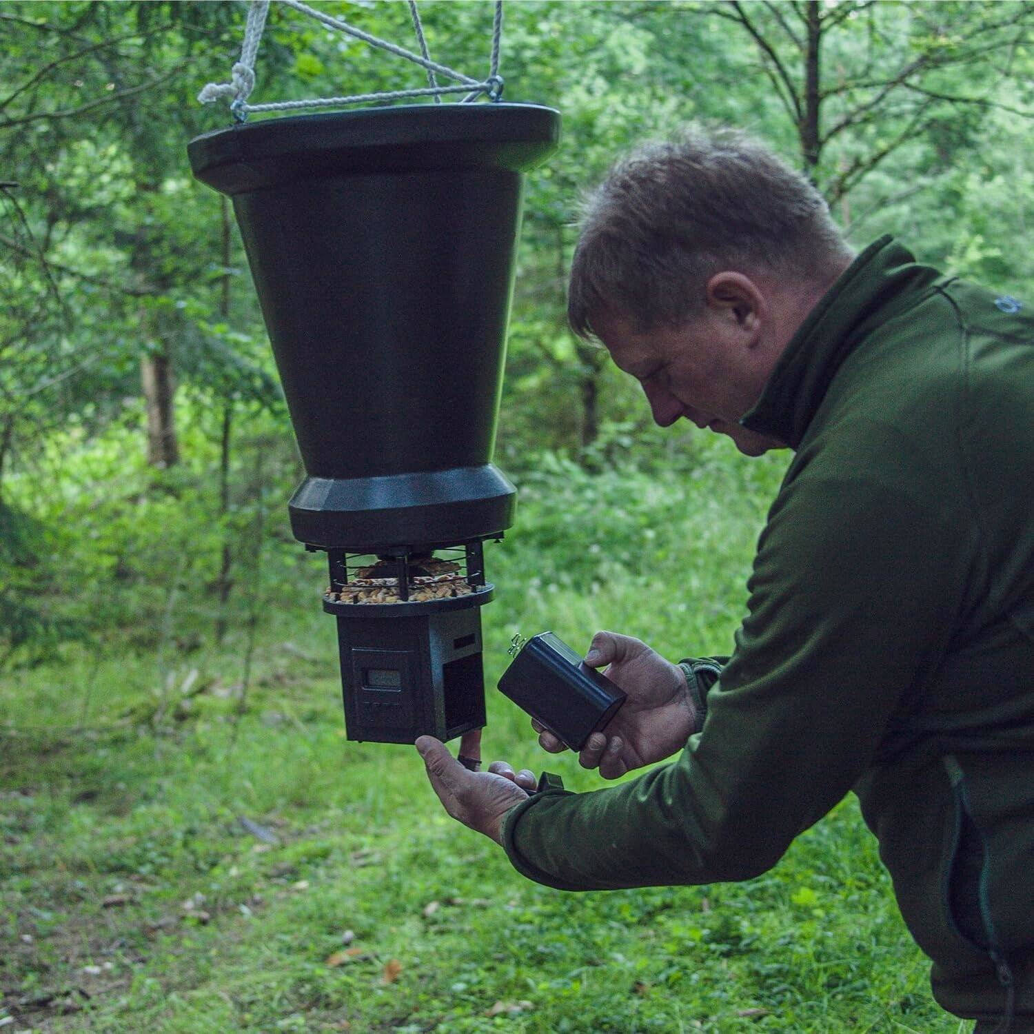Ein Mann in einer Außenumgebung richtet eine hängende Futterstelle ein, indem er Futter in sie hineingießt. Die Futterstelle ist an Bäumen aufgehängt und von Grünpflanzen umgeben.