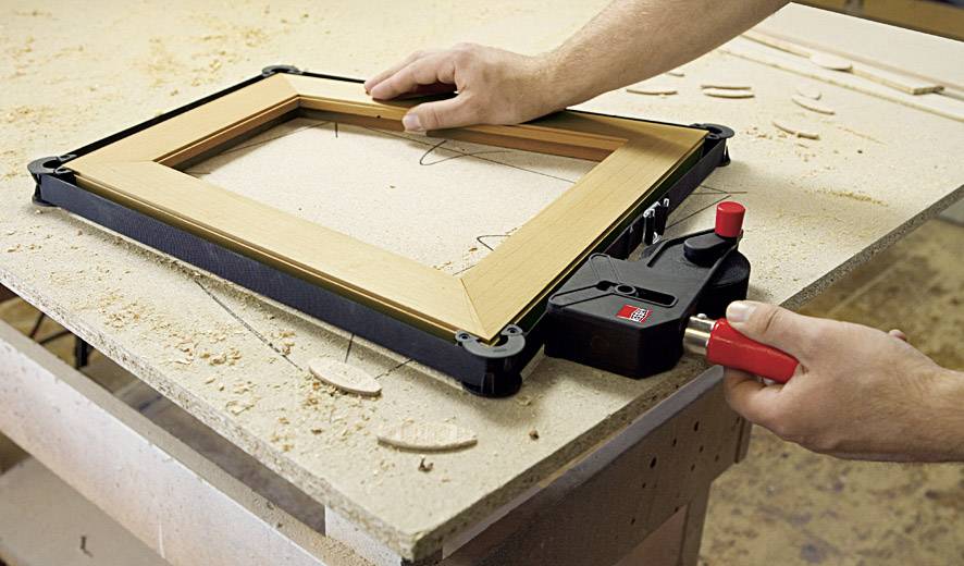 Eine Person verwendet ein Werkzeug, um einen Holzrahmen auf einer Werkbank zusammenzusetzen. Holzspäne sind um den Rahmen verteilt.