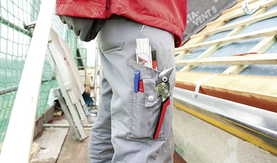 Ein Arbeiter auf einer Baustelle mit grauer Hose und roten Jacke, deren Tasche mit Werkzeugen gefüllt ist, steht neben einem Dachgerüst.