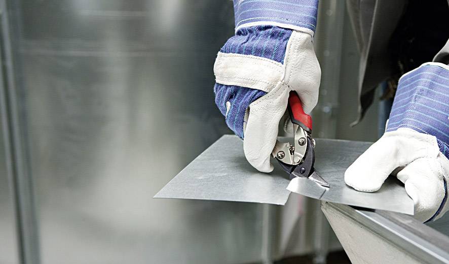 Person schneidet ein Stück Metall mit Metallschere, trägt Schutzhandschuhe. Arbeit in einer Werkstattumgebung.