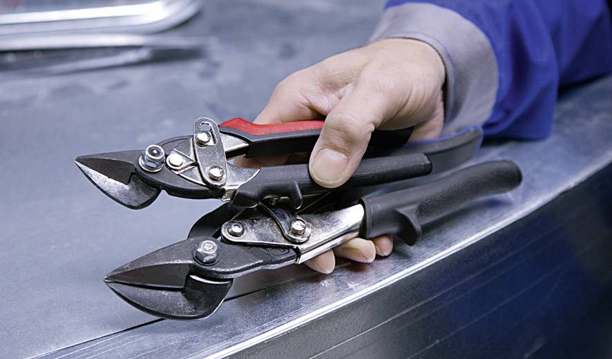 Eine Hand hält zwei Blechscheren über einer Metalloberfläche, bereit zum Schneiden.