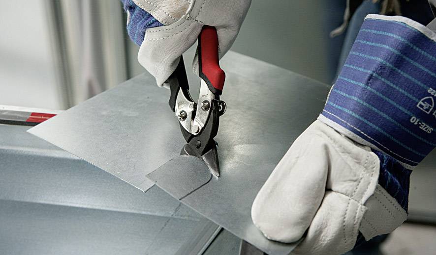 Person in Handschuhen schneidet mit einer Blechschere ein Stück Metallblech.