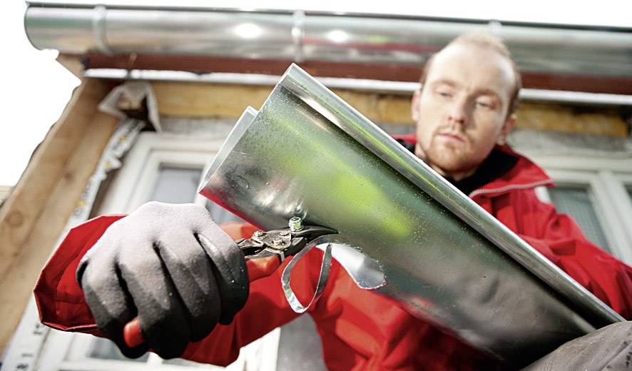 Ein Handwerker in einer roten Jacke befestigt eine metallene Regenrinne mit einer Zange an einem Haus.