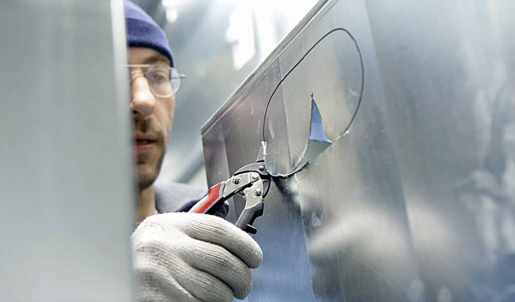 Eine Person schneidet mit einer Zange ein ovales Loch in ein Metallblech. Sie trägt Handschuhe und eine Mütze.