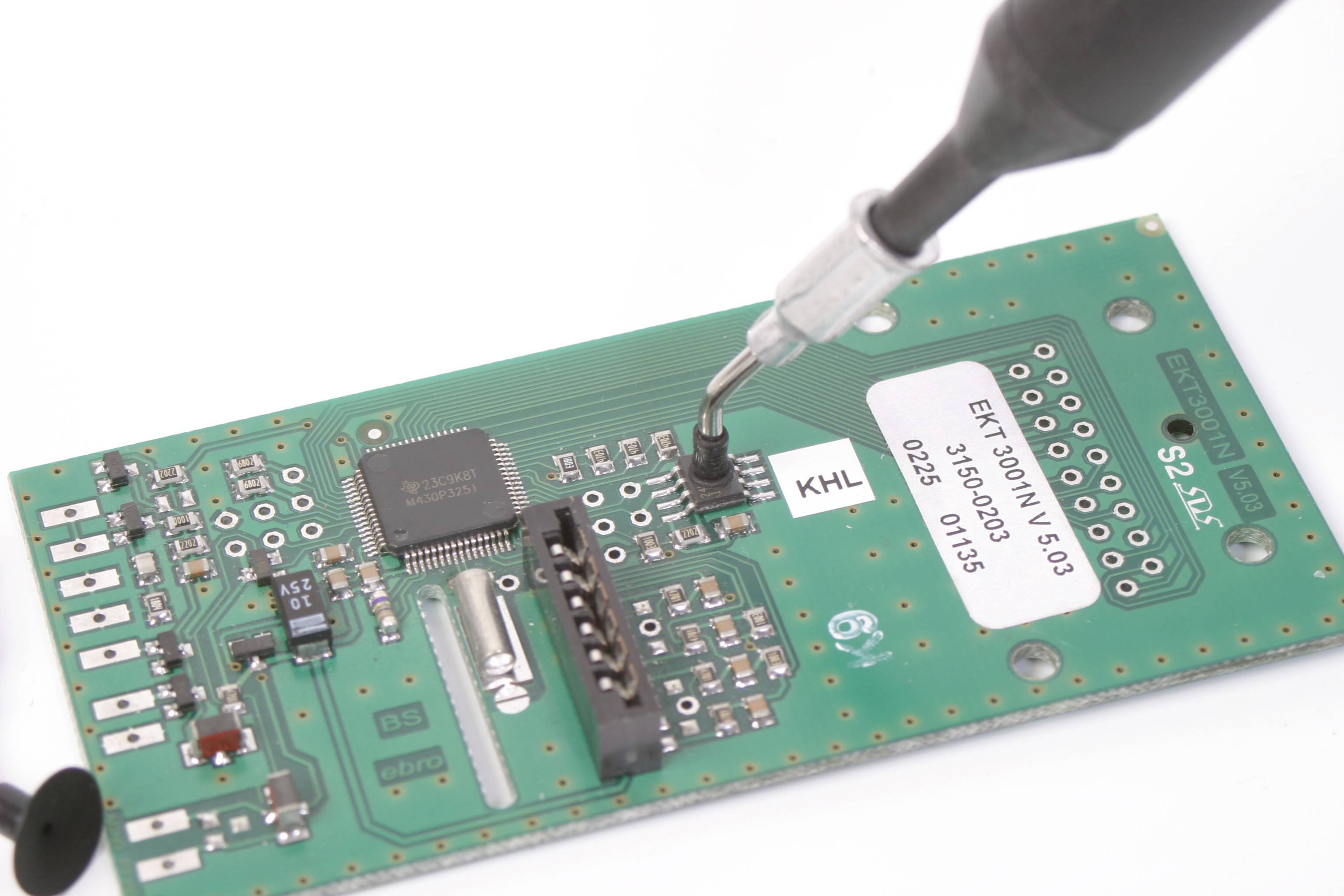 Eine Hand lötet einen grünen Schaltkreis auf einer Platine, auf dem mehrere elektronische Komponenten und silberne Anschlüsse zu sehen sind.