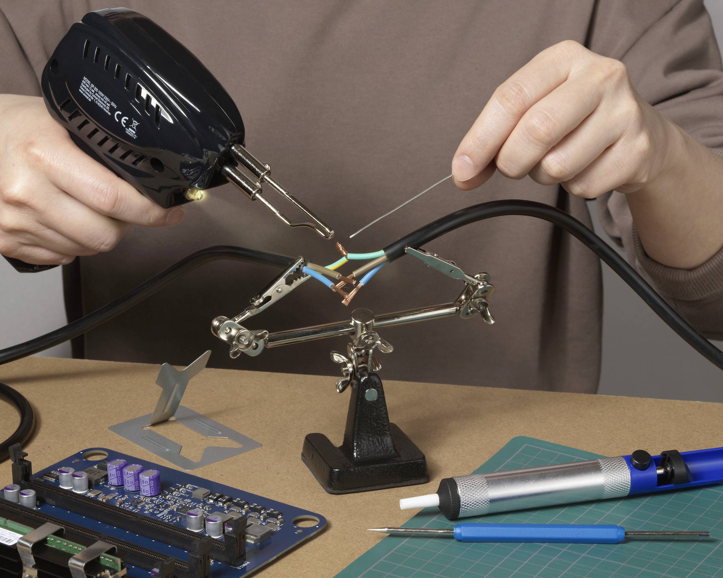 Eine Person lötet Drähte an einem schwarzen Kabel auf einem Tisch. Lötkolben und Platine sind zu sehen.