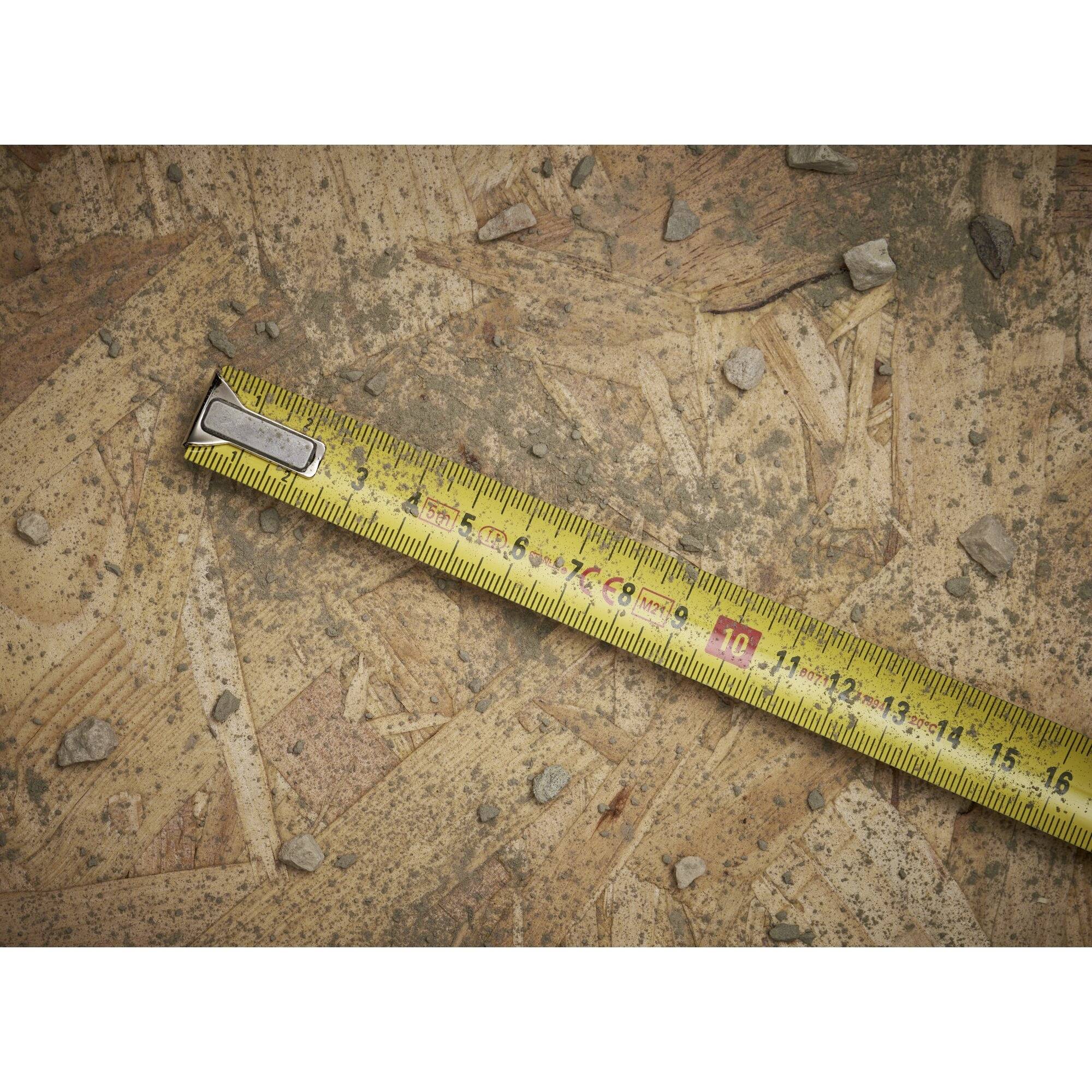 Ein gelbes Maßband liegt auf einem Boden aus Holzplanken, umgeben von kleinen Steinchen und Staub.
