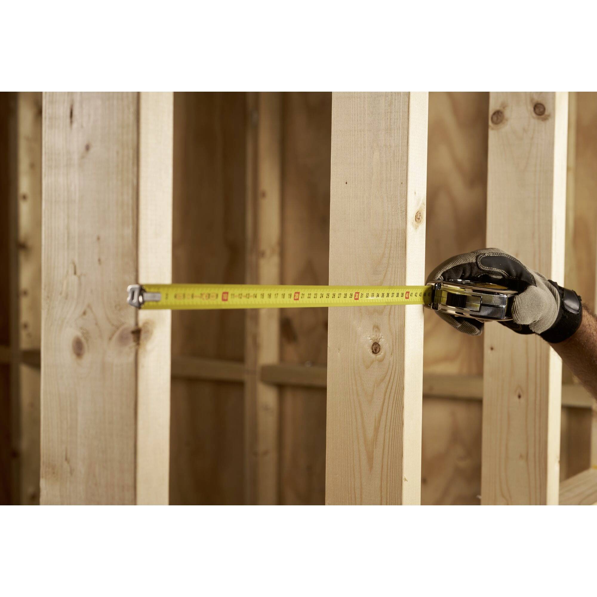 Eine Hand mit Handschuh hält ein Maßband, das Holzlatten misst. Hintergrund zeigt unvollendete Holzwand.