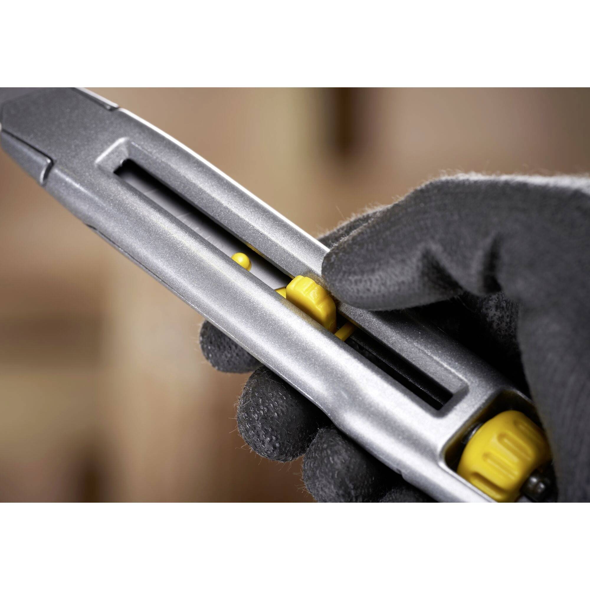 Eine mit schwarzem Handschuh geschützte Hand bedient einen silbernen Cutter mit gelben Details, schiebt die Klinge vorwärts.