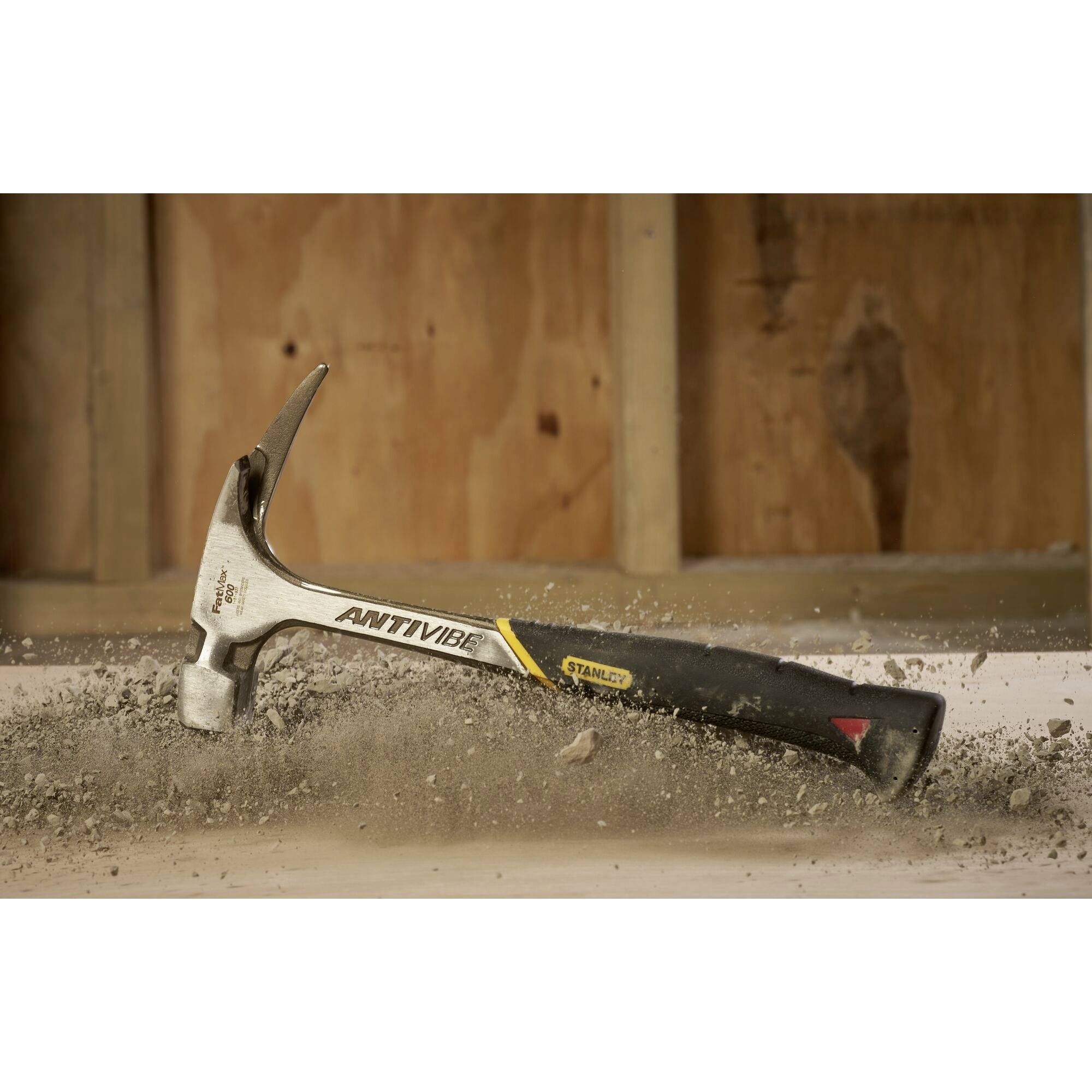 Silberner Hammer mit schwarzem Griff schlägt in Beton und erzeugt Staubwolke. Hintergrund zeigt unvollendete Holzwand.