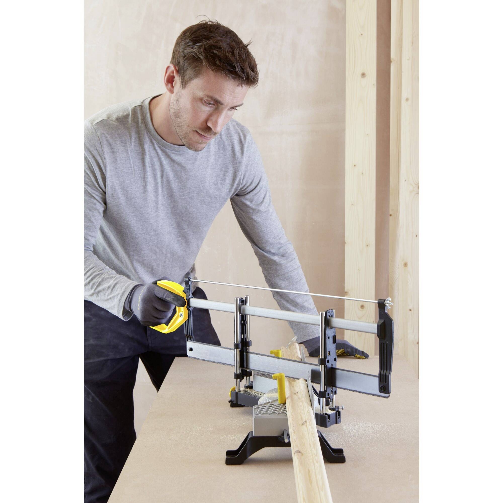 Ein Mann benutzt eine Gehrungssäge, um ein Holzstück präzise zu schneiden. Im Hintergrund sind hölzerne Strukturen sichtbar.