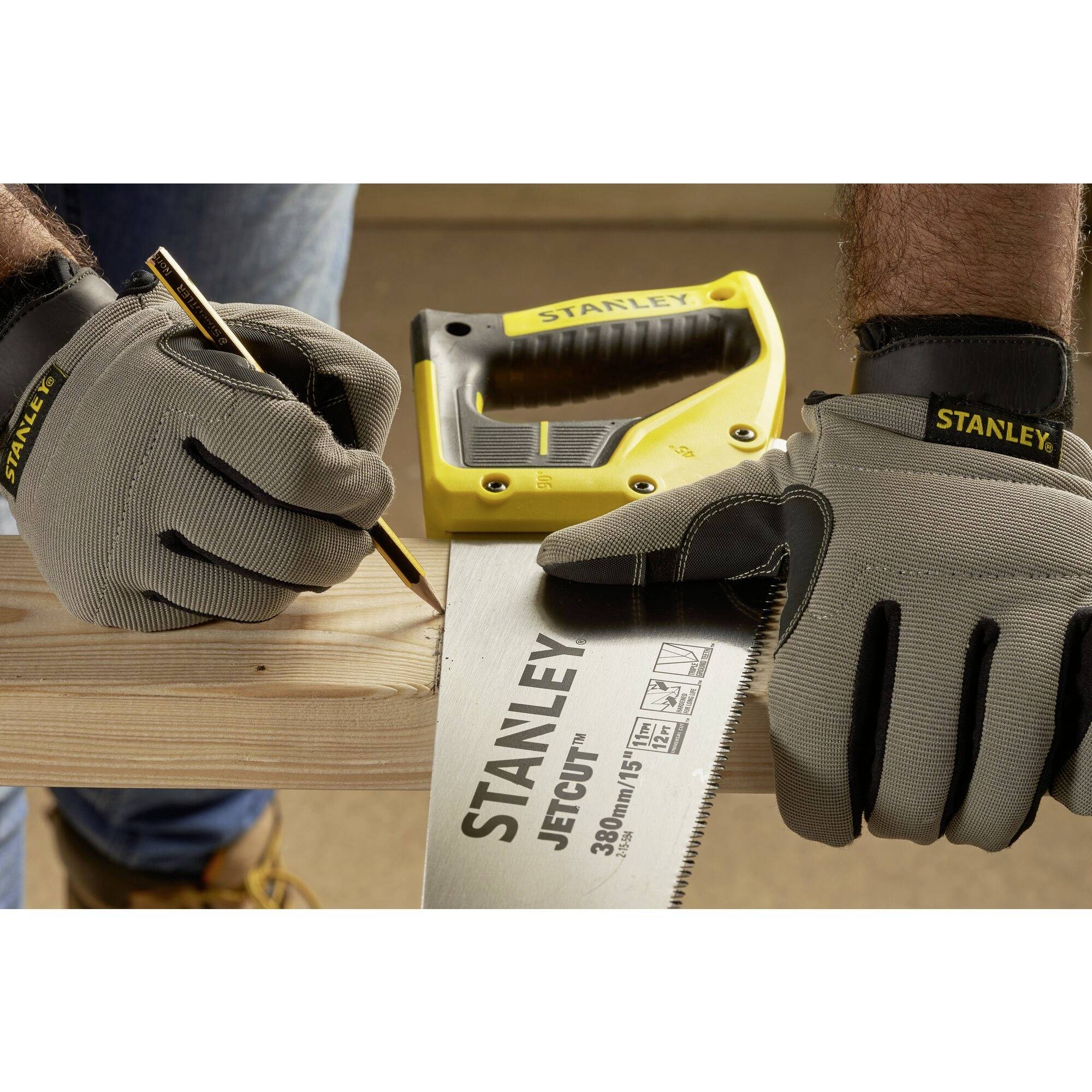 Person mit Handschuhen markiert Holz mit Bleistift, Säge und Maßband nah sichtbar, Marke 'Stanley' auf Werkzeugen.