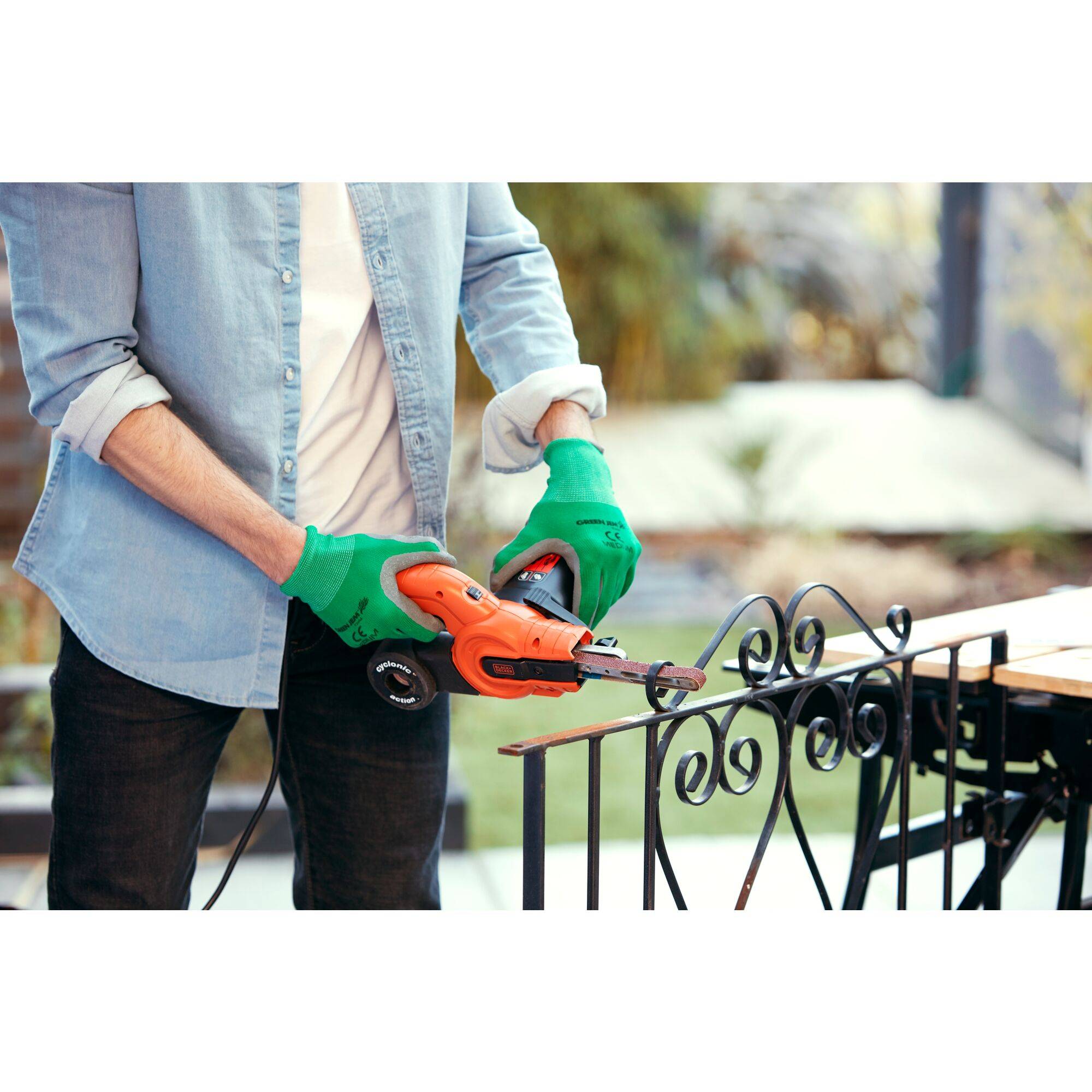 'Person mit grünen Handschuhen nutzt Bohrmaschine, um Metallgeländer zu bearbeiten. Im Hintergrund unscharfe Gartenumgebung.'