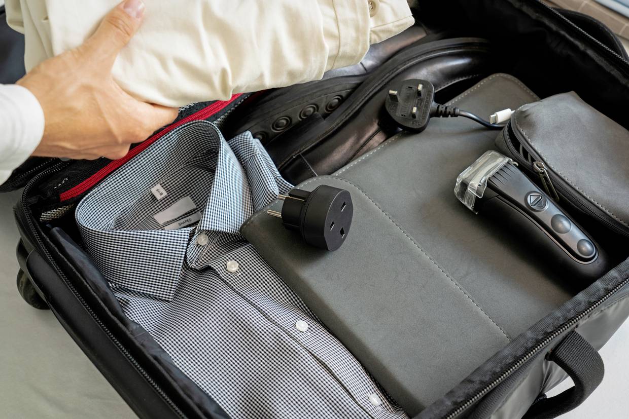 Eine offene Reisetasche zeigt eine gepackte Hemd, Schuhe, Ladegerät, Adapter und eine Person, die Hosen hineinlegt.