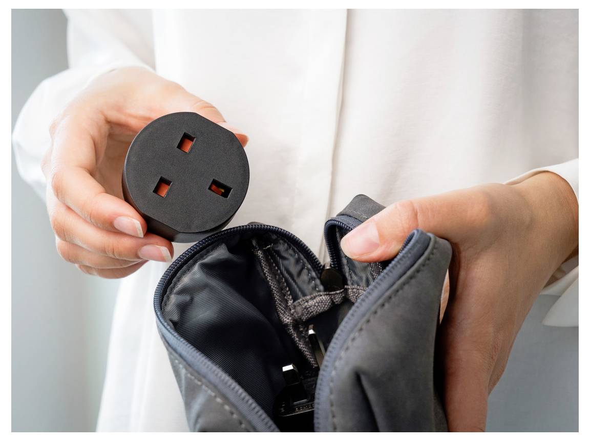 Hände halten einen runden schwarzen Stromstecker über einer offenen schwarzen Tasche und deuten darauf hin, dass ein Netzteil eingepackt oder transportiert wird.