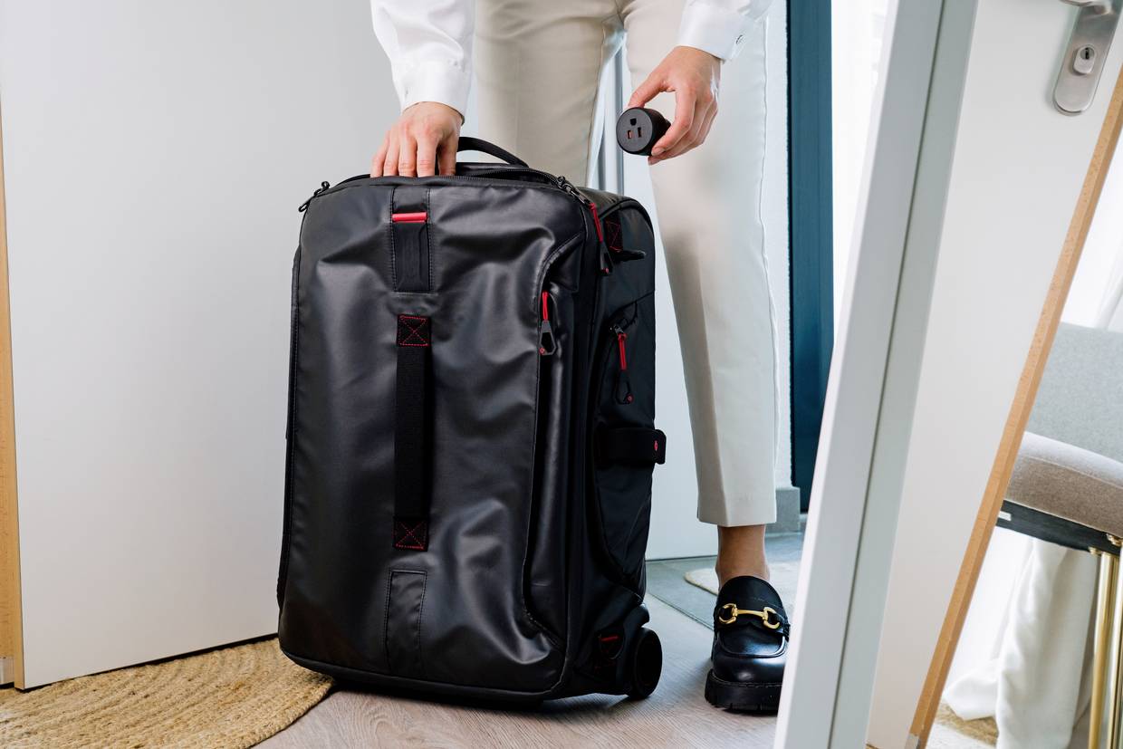 Person zieht einen schwarzen Rollkoffer durch eine Tür. Sie trägt helle Kleidung und schwarze Schuhe. Der Koffer hat rote Akzente.