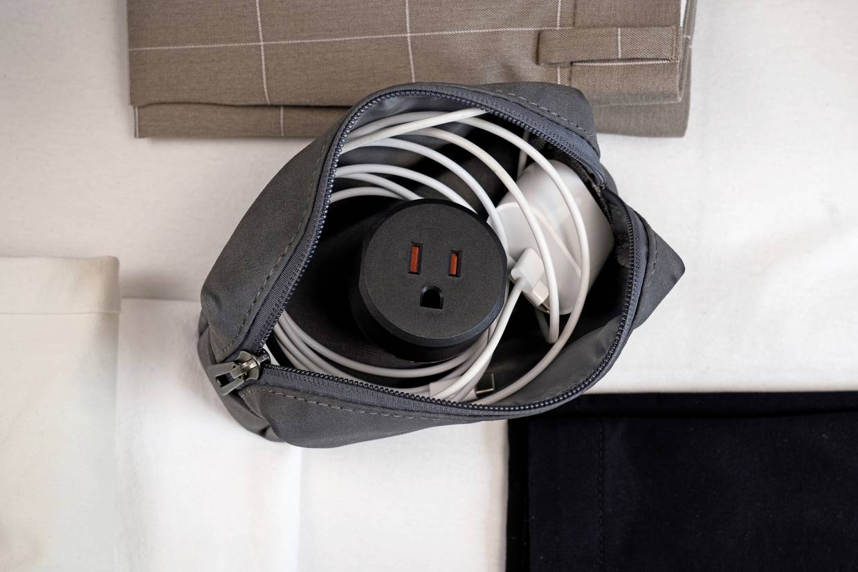 Ein graues Täschchen mit geöffnetem Reißverschluss auf einem Tisch enthält ein ordentlich aufgerolltes weißes Kabel und einen Adapter.