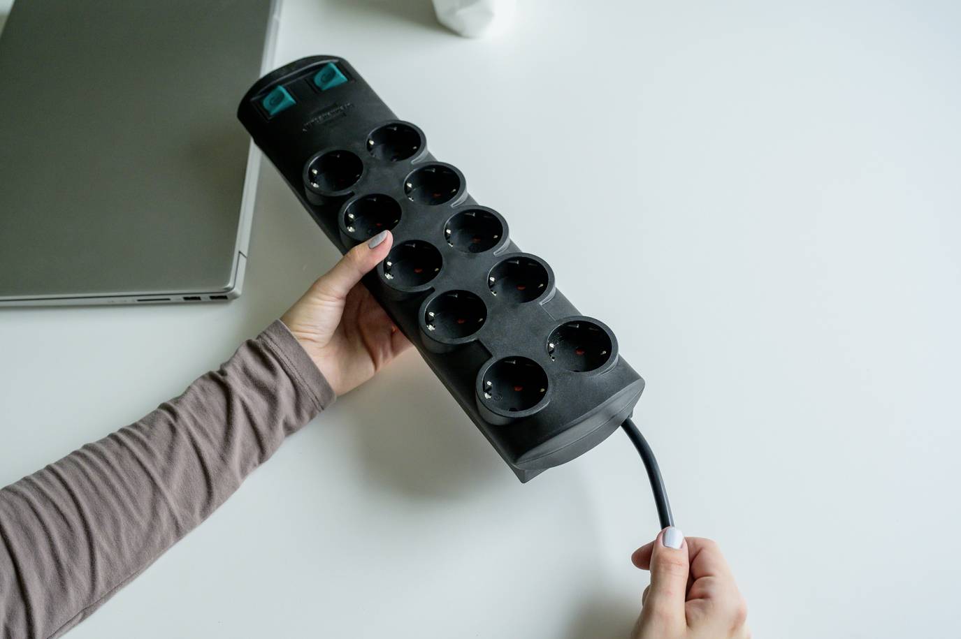 Eine Person hält eine schwarze Mehrfachsteckdose mit mehreren Steckplätzen in der Hand, auf einem Tisch neben einem Laptop.