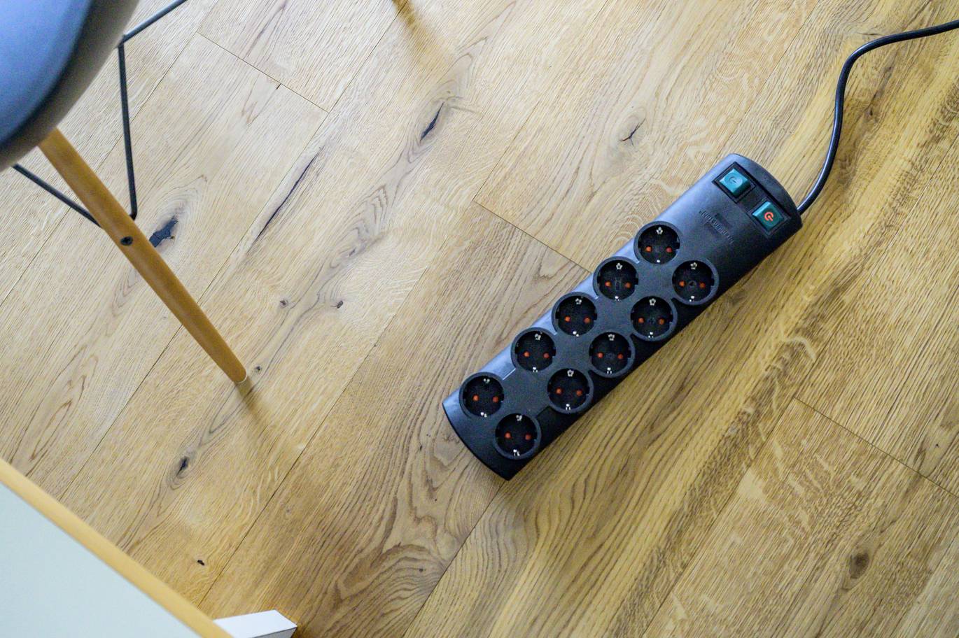 'Mehrfachsteckdose mit zehn Steckplätzen auf einem Holzboden, neben einem Stuhlbein.'