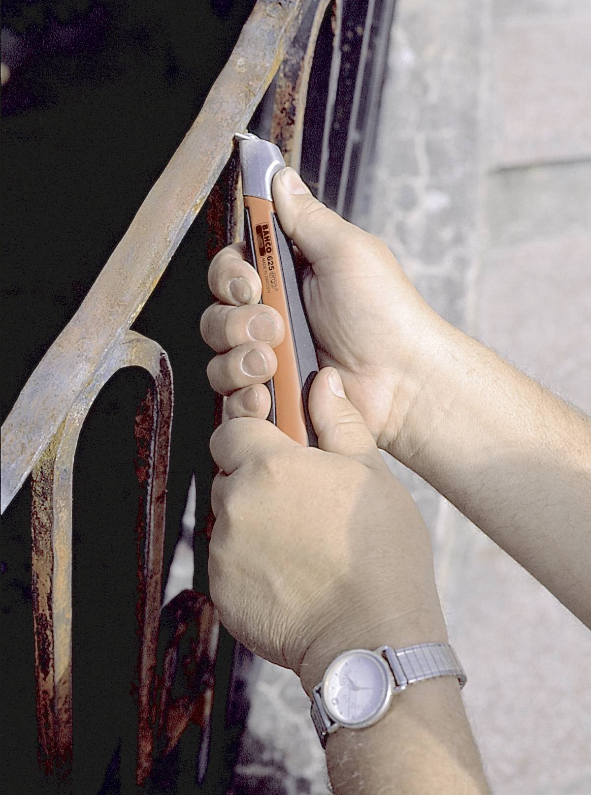 Nahaufnahme von Händen, die mit einem Schaber an einem rostigen Geländer arbeiten. Eine Armbanduhr ist am Handgelenk sichtbar.