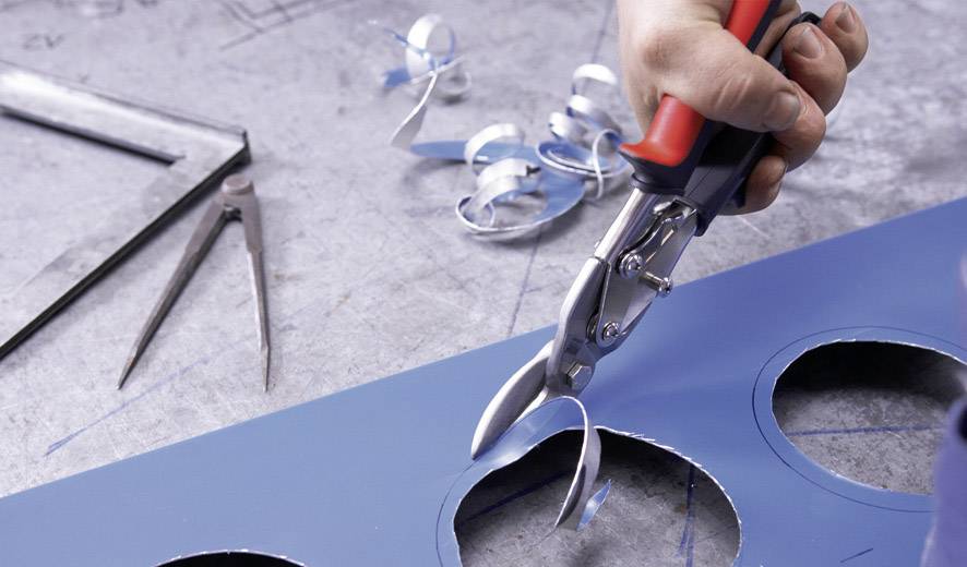 Eine Person schneidet mit einer Schere runde Löcher in ein blaues Blech. Schneidreste sind auf dem Tisch sichtbar.