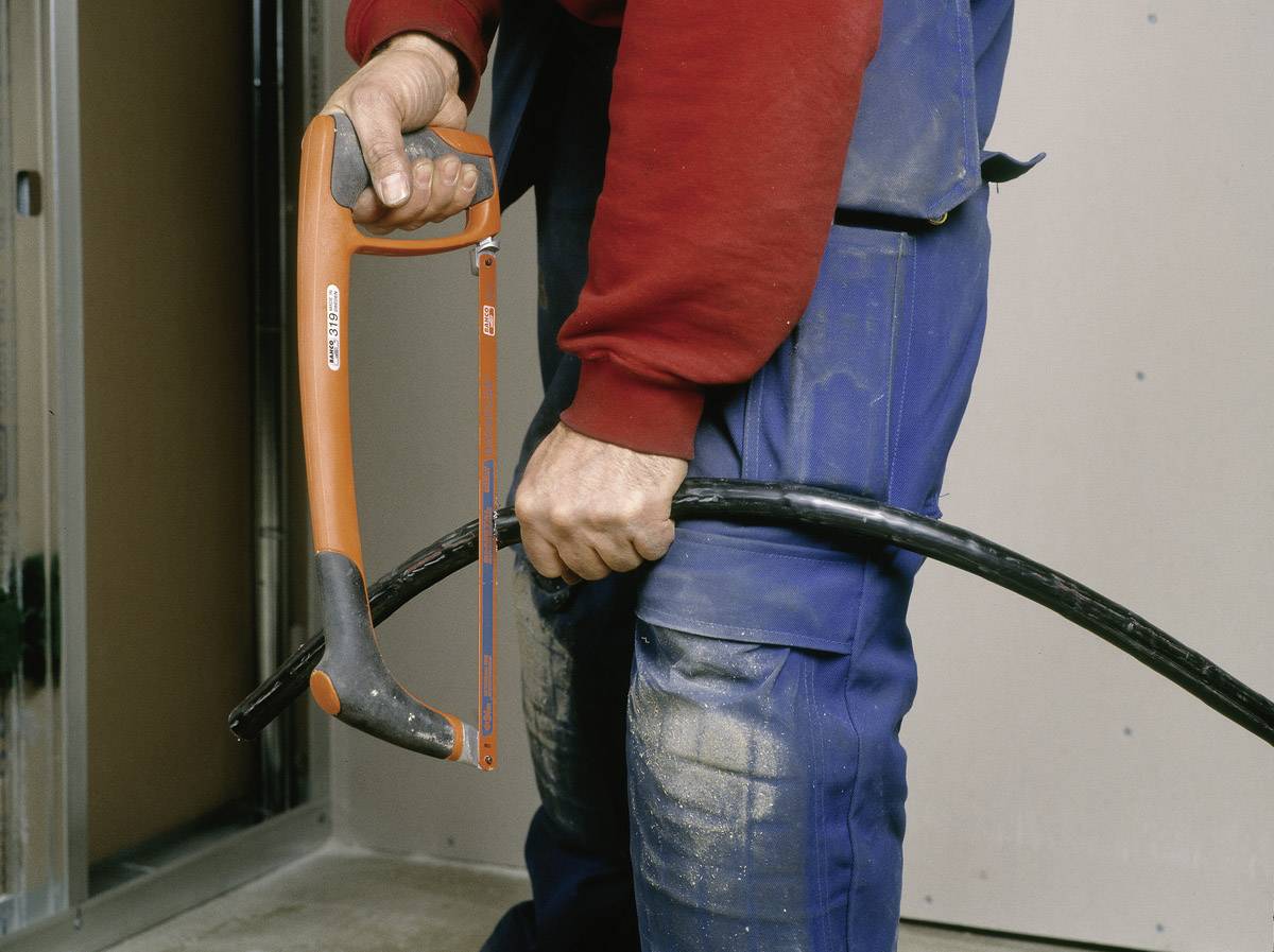 Eine Person hält eine Metallsäge und ein dickes schwarzes Kabel, bereit zum Schneiden, in einem Raum mit unfertigen Wänden.