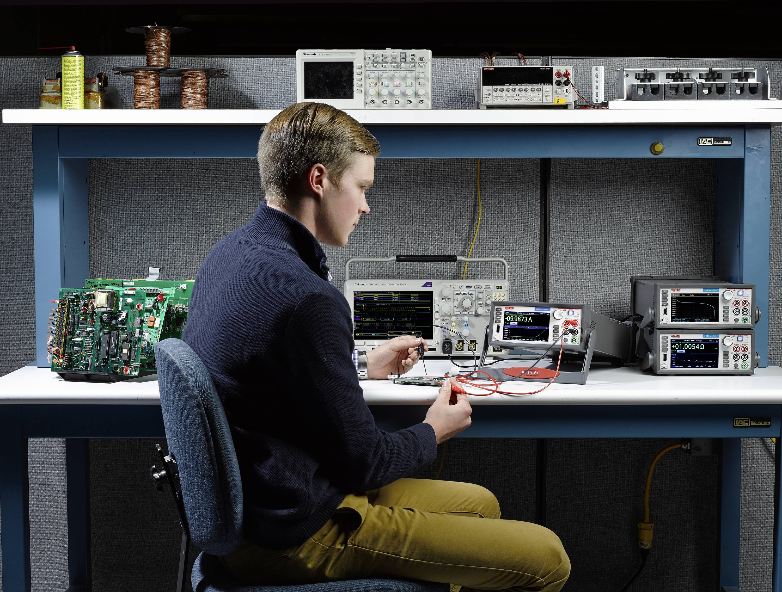 Junger Mann testet eine Platine im Labor mit Oszilloskop, Digitalmultimeter und gestapelten Labornetzgeräten.