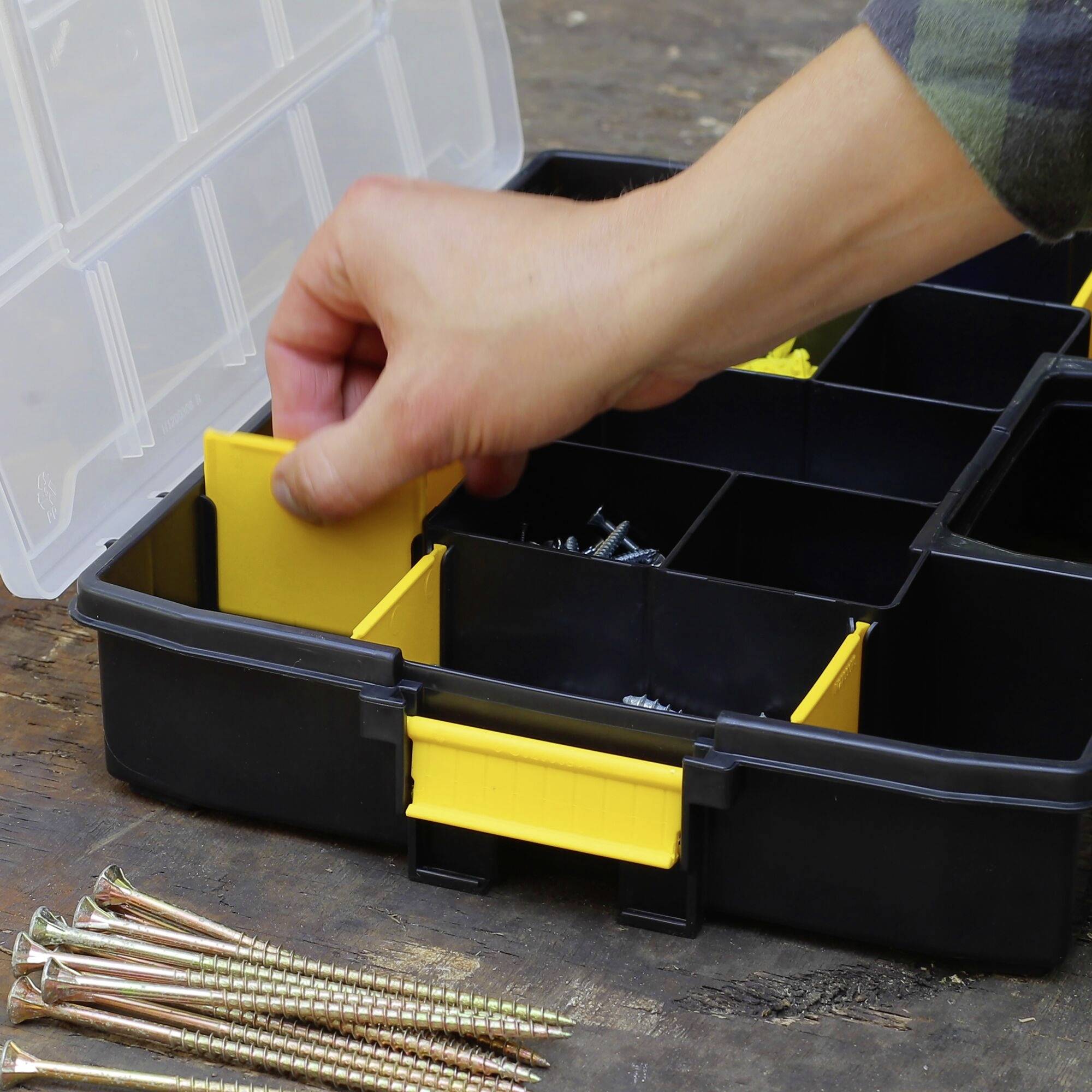 Eine Hand greift in eine offene Werkzeugbox mit mehreren Fächern, die Schrauben und Nägel enthält; einige Nägel liegen daneben.