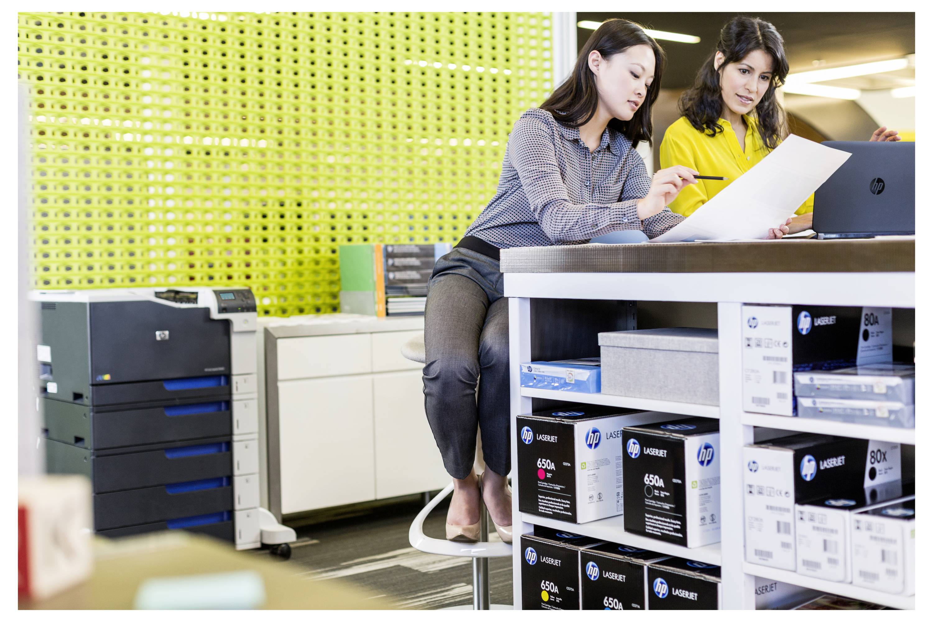 Zwei Frauen überprüfen ein Dokument an einem Schalter in einem modernen Büro- oder Einzelhandelbereich mit Regalen voller verpackter Materialien und einem Drucker in der Nähe.