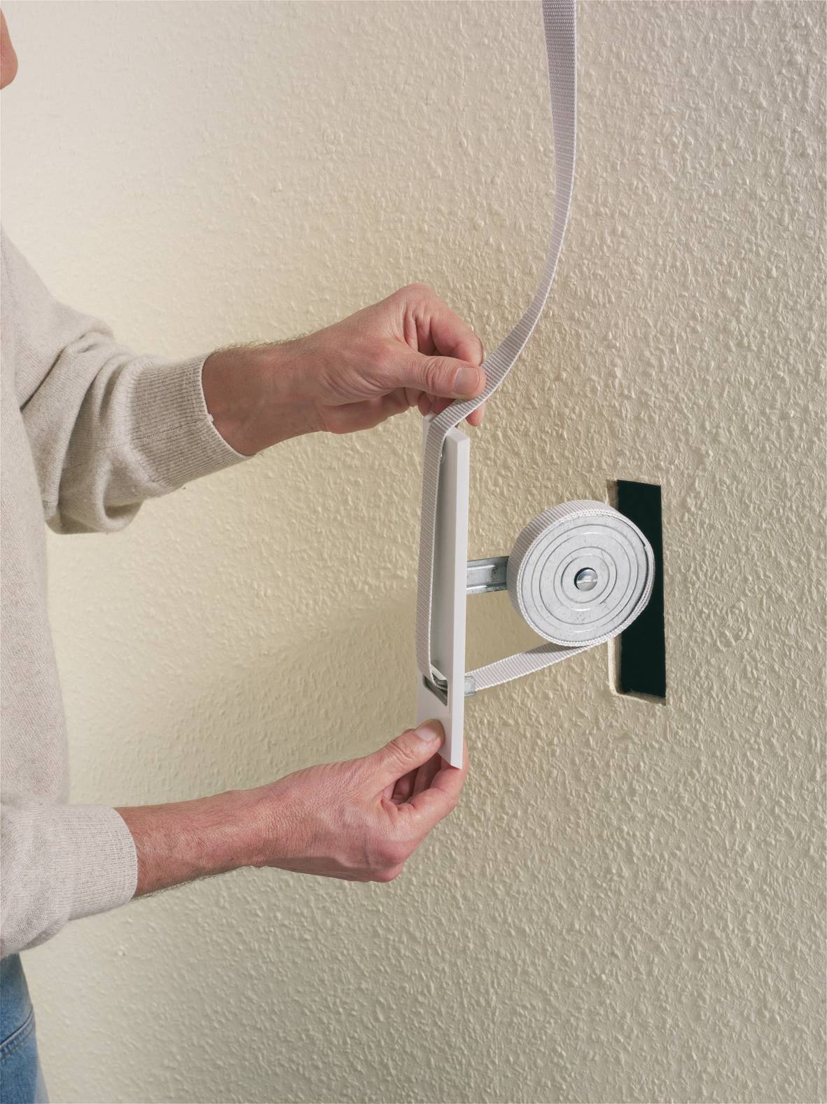 Eine Person montiert einen Rollladengurt an einer Wand. Die Hände halten den Gurtwickler und befestigen das Band am Gerät.