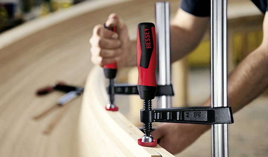 Eine Person zieht mit Schraubzwingen Holzbretter fest, um eine geformte Holzstruktur zu bauen. Werkzeugmarke 'BESSEY' gut sichtbar.