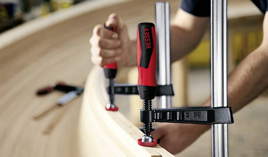 Eine Person befestigt Holzplatten mit roten Schraubzwingen an ein gebogenes Werkstück. Im Hintergrund liegen Werkzeuge.