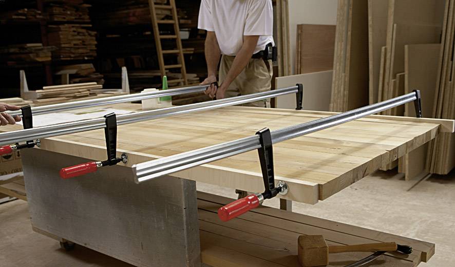 Zwei Personen arbeiten in einer Werkstatt an einem Holztisch, der mit langen Metallklemmen zur Stabilisierung fixiert ist. Im Hintergrund sind Holzmaterialien sichtbar.
