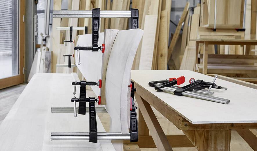 Holzbretter werden in einer Werkstatt mit Schraubzwingen fixiert, um verleimt zu werden. Im Hintergrund sind Holzbretter zu sehen.