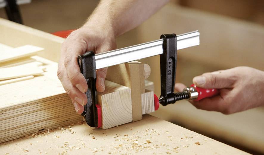 Eine Person benutzt eine Schraubzwinge, um Holzstücke auf einer Werkbank zu fixieren. Holzspäne sind auf der Oberfläche verteilt.