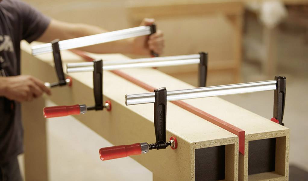 Ein Holzarbeiter befestigt zwei Holzbretter mit großen Klemmen in einer Werkstatt. Die Klemmen halten die Bretter parallel zusammen.