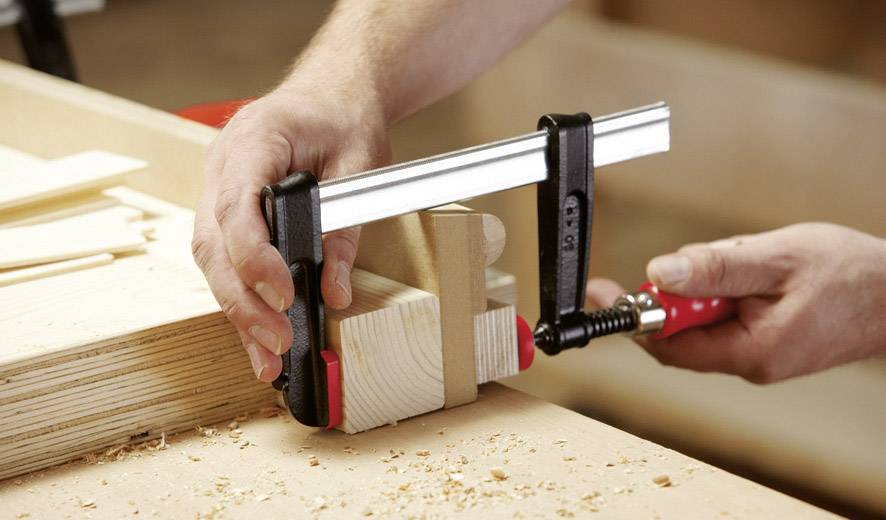 'Hand spannt Holzteile mit Schraubzwinge auf Werkbank, um sie zu fixieren. Holzspäne auf der Oberfläche zeigen kürzliches Sägen.'