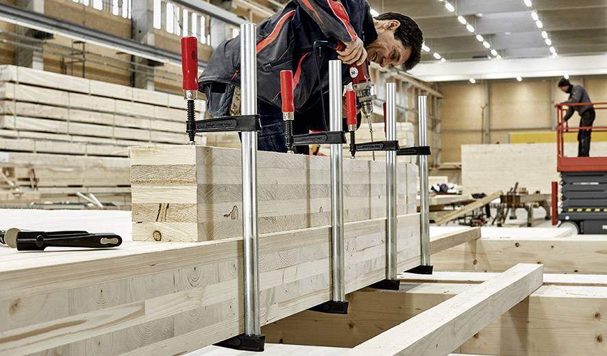 'Eine Person befestigt Holzplatten mit großen Klammern in einer Werkstatt. Der Raum ist gefüllt mit Holzbrettern und Werkzeugen.'