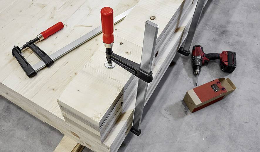 Holzbretter werden mit Schraubzwingen stabilisiert; im Hintergrund Bohrmaschine und Schraubkästen – Szene zeigt Handwerksarbeiten.