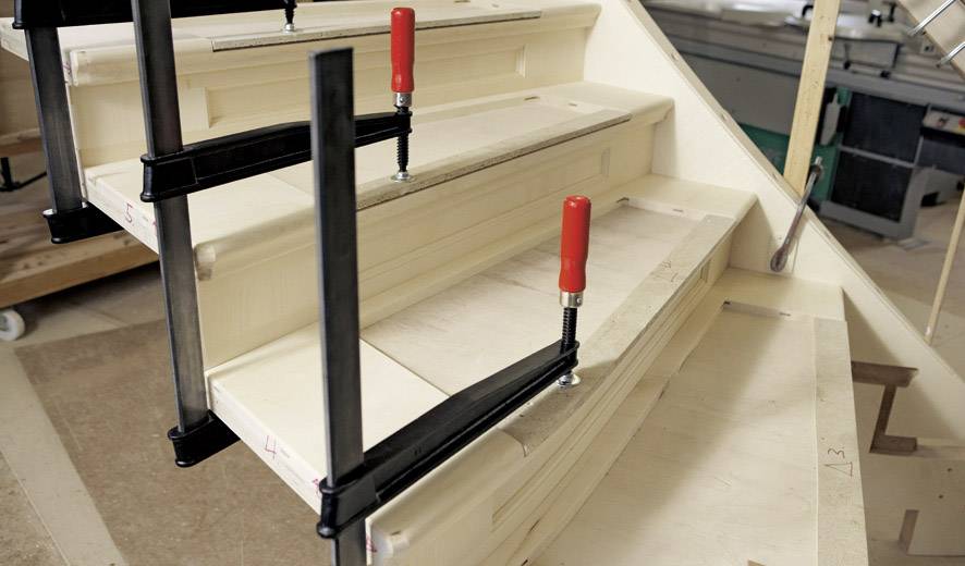 Eine halbgebaute Holztreppe wird mit roten Zwingen fixiert. Handwerksarbeiten in einer Werkstatt.