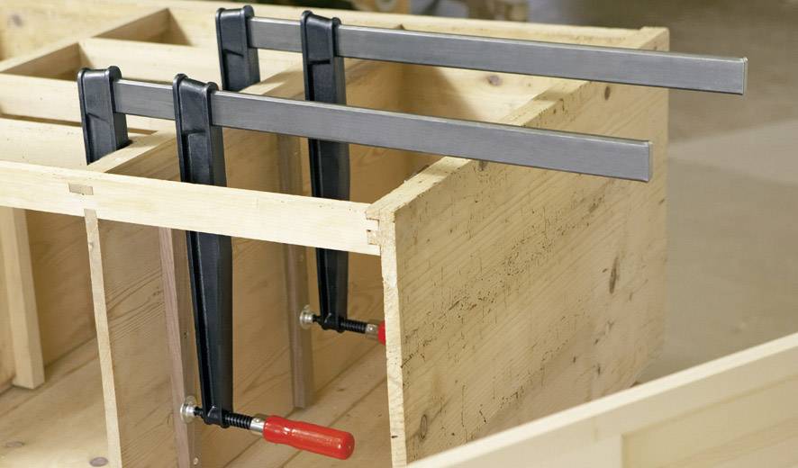 Drei schwarze Schraubzwingen halten Holzplatten zusammen. Werkstattumgebung.