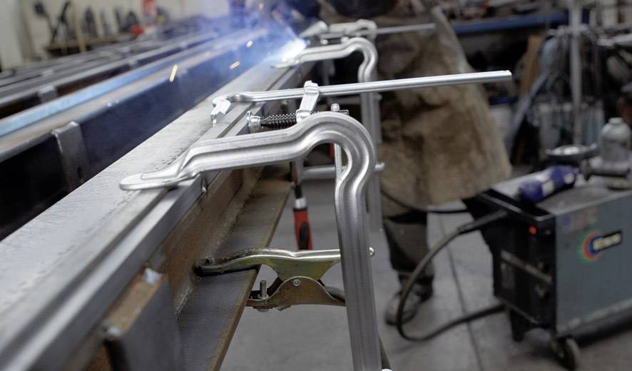 Eine Person schweißt Metall in einer Werkstatt. Im Vordergrund sind Zwingen zu sehen, die Metallteile fixieren. Funken sprühen.