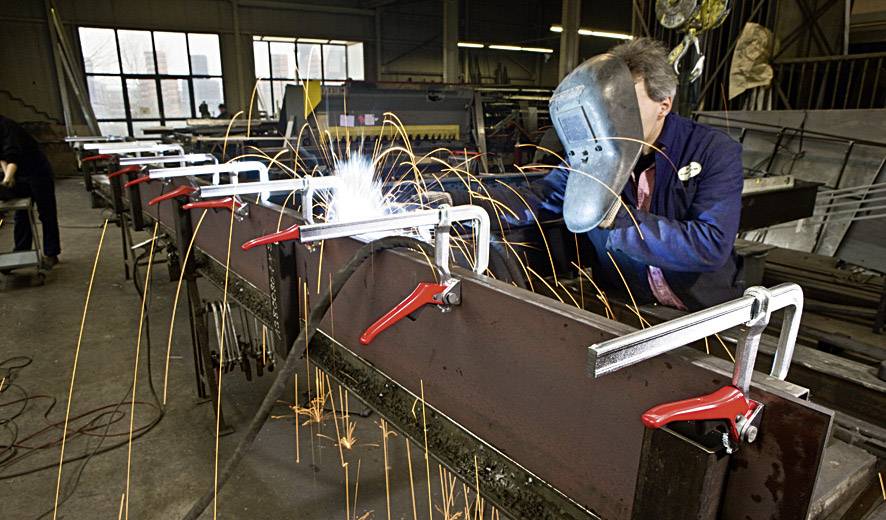 Eine Person schweißt an einer Metallkonstruktion in einer Fabrikhalle. Funken sprühen, mehrere Werkstücke sind mit Klemmen fixiert.