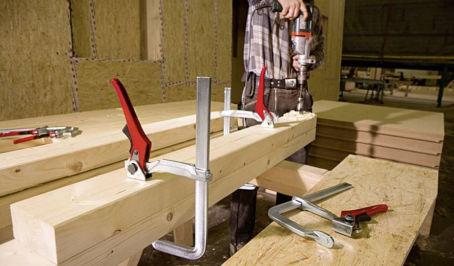 Eine Person verwendet eine Bohrmaschine, um Löcher in Holz zu bohren. Holzplatten und Klemmen sind auf einem Tisch sichtbar.