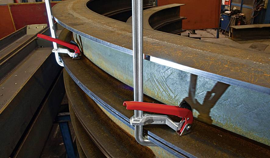 'Zwei große Stahlplatten werden mit roten Schraubzwingen an einer Werkbank befestigt, um gebogene Stahlträger zu sichern.'