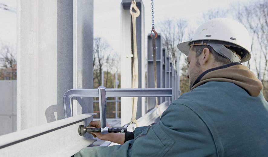 Ein Bauarbeiter mit Schutzhelm befestigt Metallteile auf einer Baustelle. Im Hintergrund sind Gerüste und Bäume zu sehen.